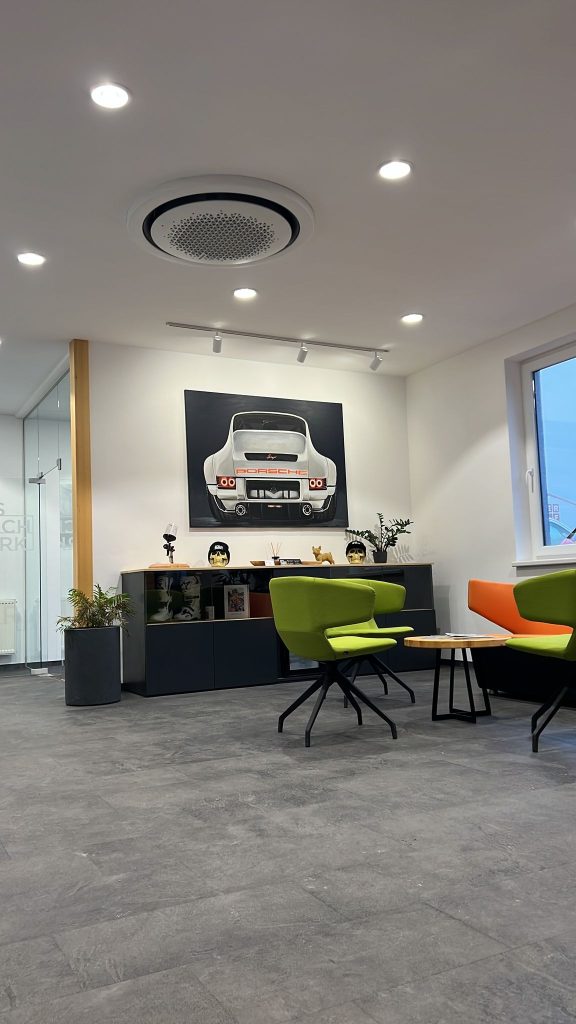 Design trifft Komfort – Samsung 360° Klimagerät im modernen Wartebereich | Klima-Expert Runder Samsung 360° Deckenluftauslass in einem stilvoll eingerichteten Raum mit Porsche-Bild